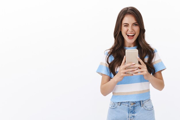 Cheeky stylish modern woman taking selfie in mirror, winking herself ambitious and flirty show tongue, smiling delighted, try new smartphone camera, photographing near white wall