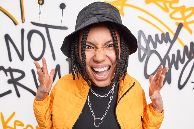 Cheeky woman with dreadlocks raises hands exclaims angrily wears fashionable clothes poses near graffiti wall at street belongs to hooligan gang