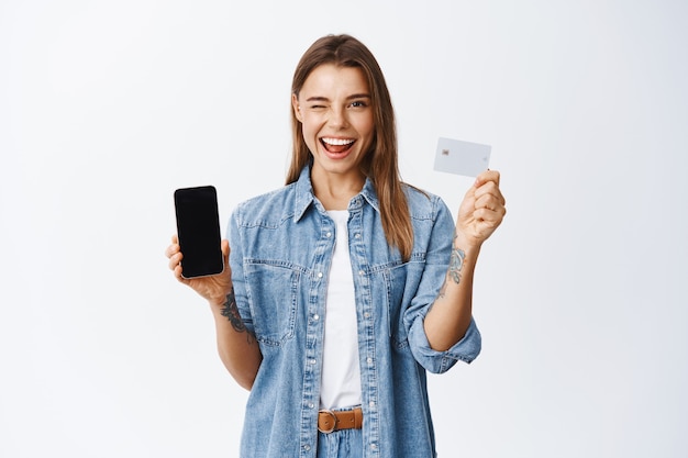 Cheeky young woman winking at you, recommending mobile banking app, showing empty smartphone screen and plastic credit card, white wall