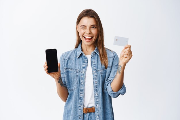 Нахальная молодая женщина, подмигивающая вам, рекомендует приложение для мобильного банкинга, показывает пустой экран смартфона и пластиковую кредитную карту, белая стена