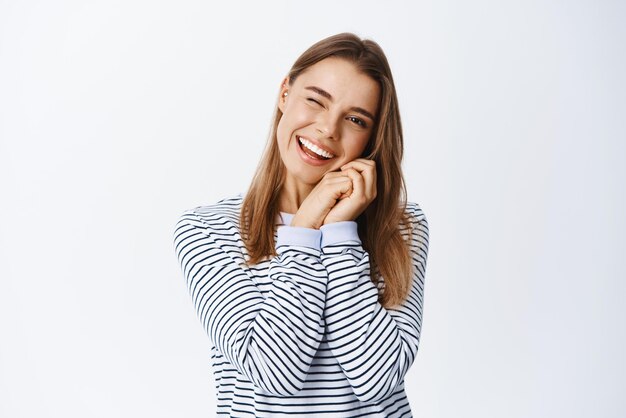 Cheeky young woman with blond hair holding hands near natural face with light make up winking and smiling at camera standing over white background