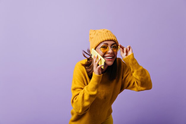 Cheerful 21 year old girl with auburn hair has funny, interesting conversation by phone. Laughing woman looking through yellow sunglasses with interest posing for portrait