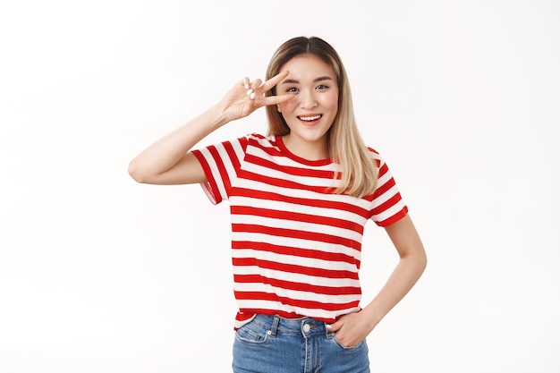 Cheerful active lively charming blond asian girl having perfect energized day show victory gesture eye disco sign smiling broadly toothy positive grin standing white wall