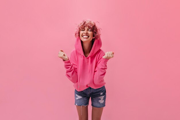 분홍색 까마귀와 데님 바지를 입은 쾌활한 활동적인 마른 소녀가 분홍색 배경에서 웃고 있는 외진 행복한 곱슬머리 여성에게 활짝 웃고 있습니다.