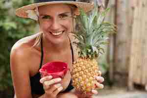 Foto gratuita la donna adorabile allegra in cappello di paglia gode delle vacanze estive sulla spiaggia tropicale, tiene l'ananas esotico e la frutta del drago