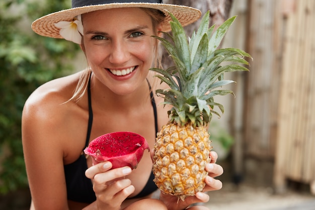 Cheerful adorable woman in straw hat enjoys summer vacation on tropical beach, holds exotic pineapple and dragon fruit