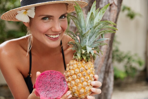 Cheerful adorable woman in straw hat enjoys summer vacation on tropical beach, holds exotic pineapple and dragon fruit