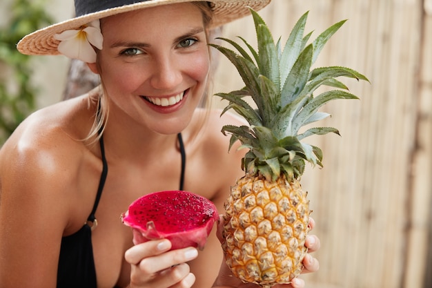 밀짚 모자에 쾌활한 사랑스러운 여자는 열대 해변에서 여름 휴가를 즐기고, 이국적인 파인애플과 용 과일을 보유