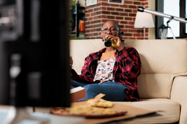 Cheerful adult drinking alcoholic beer from bottle on couch, watching movie or film on television channel. Eating fast food meal from delivery package and enjoying beverage in living room.