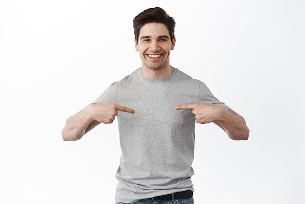 흰색 배경 위에 서 있는 배너 회사 홍보 문구를 가리키는 귀하의 로고가 표시된 센터 티셔츠를 가리키며 웃고 있는 쾌활한 성인 남자