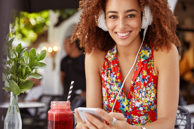 Cheerful African American female with curly hair listens cool composition in headphones, enjoys good relaxation and summer rest in sidewalk restaurant with cocktail.