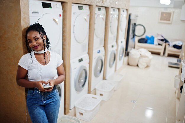 Веселая африканская американка возле стиральной машины слушает музыку в наушниках с мобильного телефона в прачечной самообслуживания