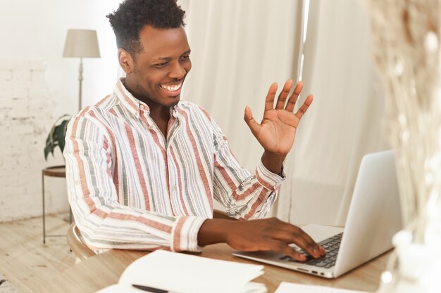 광범위 하 게 웃 고 오픈 노트북 앞에 앉아 쾌활 한 아프리카 학생, wifi를 사용 하여 온라인 공부하는 동안 교사를 인사.