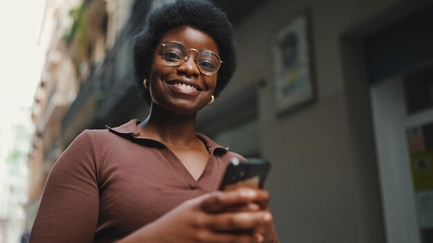 Cheerful African woman in glasses holding smartphone on the stre