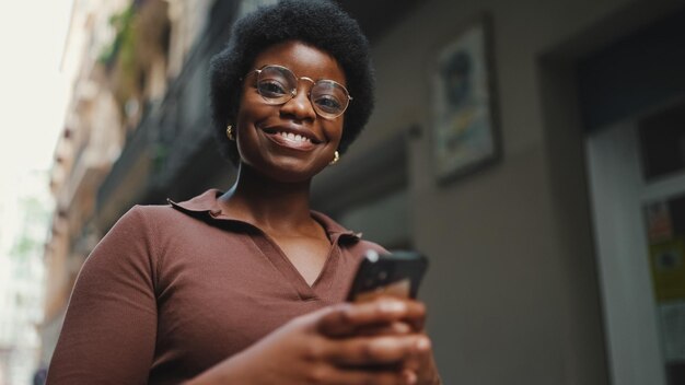 通りにスマートフォンを保持している眼鏡の陽気なアフリカの女性