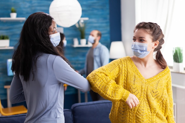 Cheerful african woman and her friend touching elbow keeping social distancing while greating each other wearing face mask, to prevent coronavirus spread in the course of global pandemic in living roo