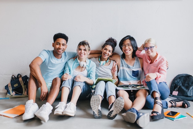 Free photo cheerful african young man in white sneakers embracing his university friends sitting on the floor with backpack. indoor portrait of smiling students resting after classes and posing with pleasure.