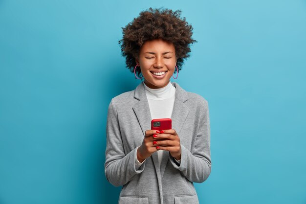 陽気なアフリカ系アメリカ人の女性ブロガーは、オンラインで画像を投稿し、電話で仕事をし、インターネットで面白いビデオを笑い、楽しんで目を閉じ、灰色のジャケットを着ています。