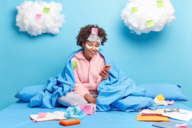 Cheerful Afro American girl works from home prepares project while being on quarantine surfs internet via smartphone