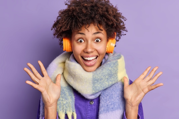 Cheerful Afro American woman looks with very happy excited expression raises hands enjoys listening favorite music via stereo headphones wears warm scarf around neck.