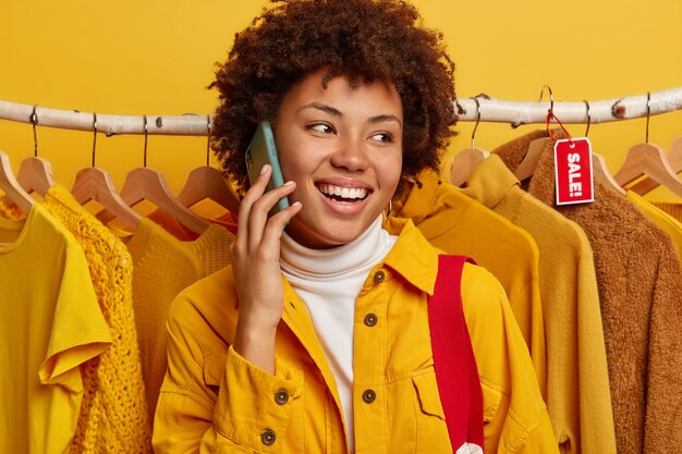 Cheerful Afro American woman talks on cell phone, checks out some clothes at store, poses over clothing rack, sharaes what sales are in shop