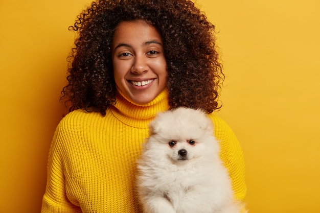 Жизнерадостная афроамериканка с зубастой улыбкой, держит белого шпица, работает волонтером, находит приют для животных, носит желтый свитер.