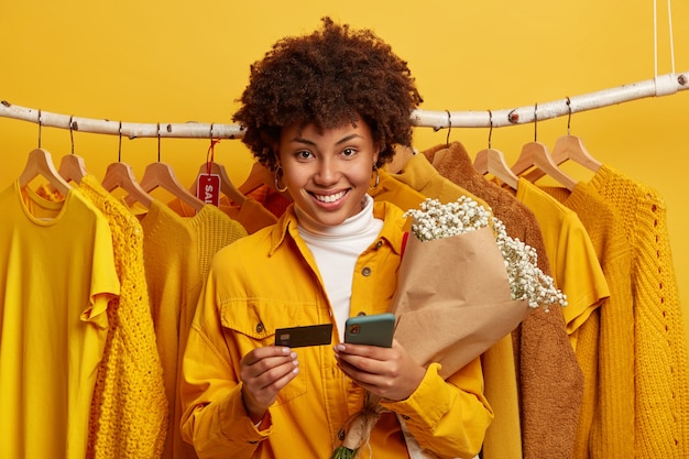 Cheerful Afro woman holds mobile, credit card and bouquet, stands against clothes rail