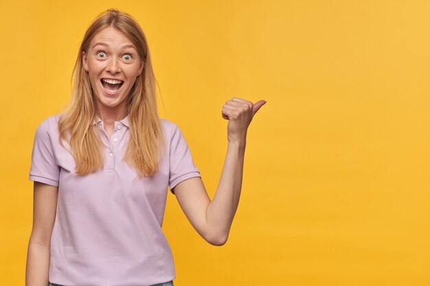 Cheerful amazed woman with freckles in lavender tshirt feels excited and pointing away to the side at copyspace on yellow