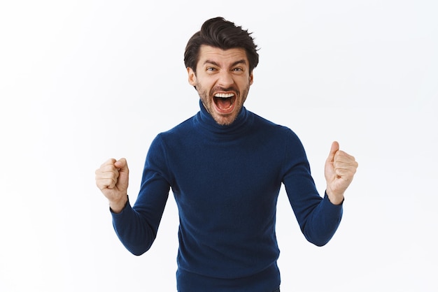 Cheerful ambitious goodlooking bearded businessman in high neck sweater yelling from happiness rooting for team fist pump and stare camera accomplished winning competition achieve goal