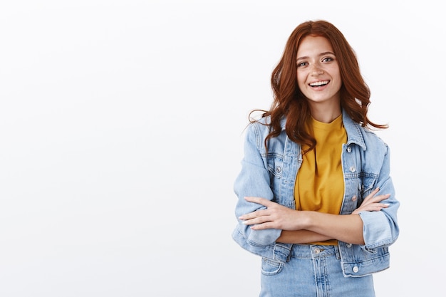 Allegra ambiziosa donna rossa in giacca di jeans incrocia le braccia in posa sicura, sorridente compiuta e sicura di sé, muro bianco in piedi