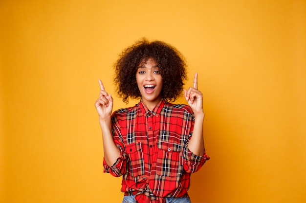 Cheerful american black woman in red  shirt looking upward and pointing fingers up on copy space isolated over orange background.