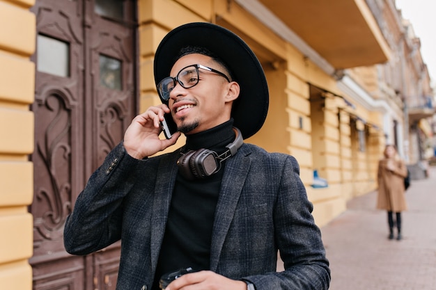 오래 된 건물 근처에 서있는 동안 전화 통화하는 유행 회색 복장에 쾌활 한 미국 사람. 누군가 전화하고 웃고 열정적 인 아프리카 남자.