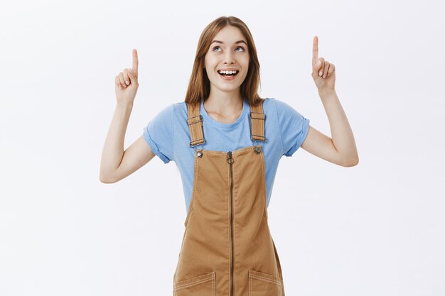 Cheerful and amused girl pointing fingers up at logo or advertisement