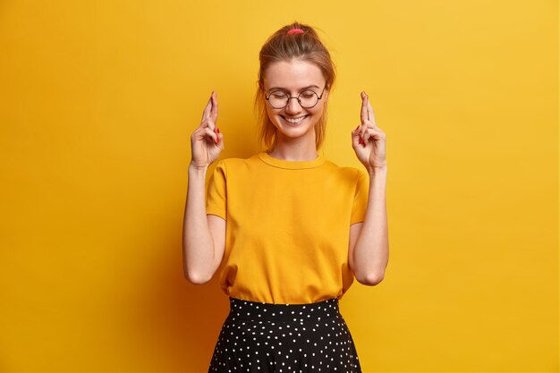 Cheerful amused millennial girl crosses fingers hopes dreams come true or wish fulfilled smiles pleasantly receives good news wears round transparent glasses t shirt and skirt isolated on yellow wall