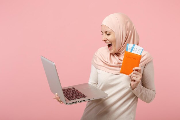 히잡 가벼운 옷을 입은 쾌활한 아라비아 이슬람 여성이 분홍색 배경에 고립된 포즈를 취하고 있습니다. 사람들이 종교 이슬람 생활 방식 개념입니다. 복사 공간을 비웃습니다. 노트북 pc 컴퓨터, 여권, 티켓을 들고 있습니다.