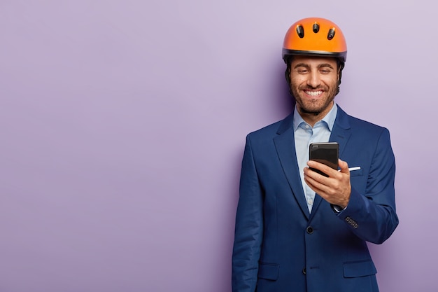 Cheerful architect wears orange hardhat, formal suit, has spare time during work break, gets message on smartphone, happy to receive salary