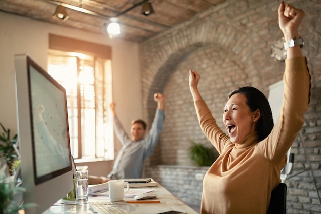 Donna d'affari asiatica allegra con le braccia alzate per celebrare la ricezione di buone notizie sul computer in ufficio