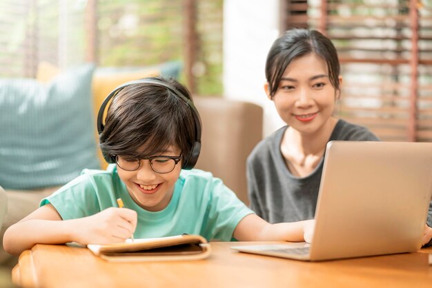 Cheerful asian child boy online lesson study from home conceptasian child hand up for answer question from online teacher asian boy sit with mom and dad learn from laptop at living area at home