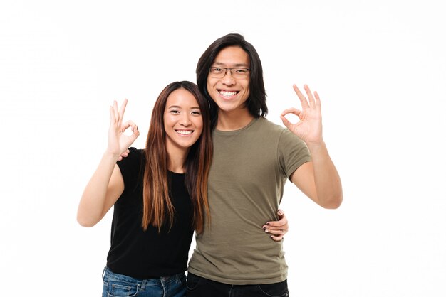Cheerful asian couple showing OK gesture while hugging each other, looking at camera