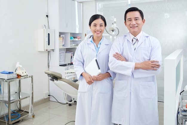 Cheerful asian dentists posing in treatment room in clinic in front of equipment