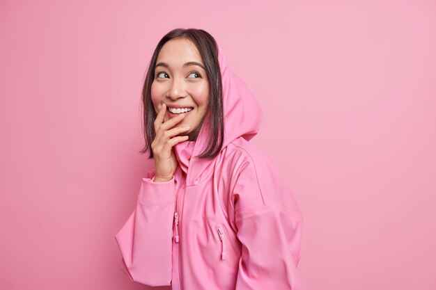 Cheerful Asian female teenager gazes aside daydreams and remembers lovely moment smiles broadly dressed in hoodie expresses positive candid emotions isolated over pink wall. Youth lifestyle