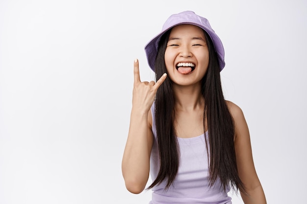 La ragazza asiatica allegra mostra la lingua e il gesto di corna di metallo pesante rock n roll godendo l'evento divertendosi in estate in piedi su sfondo bianco