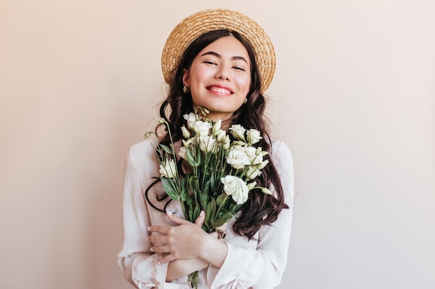Веселая азиатка в соломенной шляпе с цветами в руках Молодая женщина с белыми эустомами, выражающая счастье