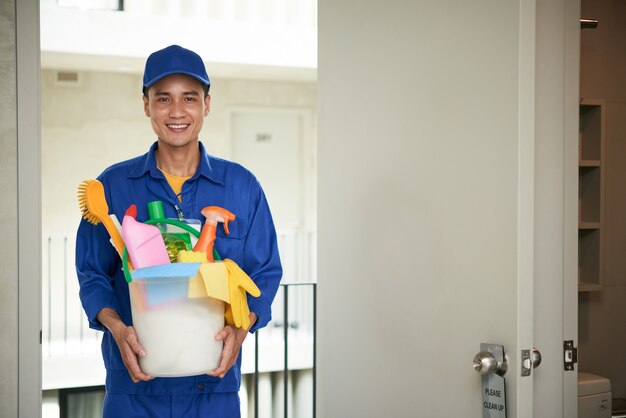 Cheerful Asian male janitor walking into hotel room, carrying supplies in bucket