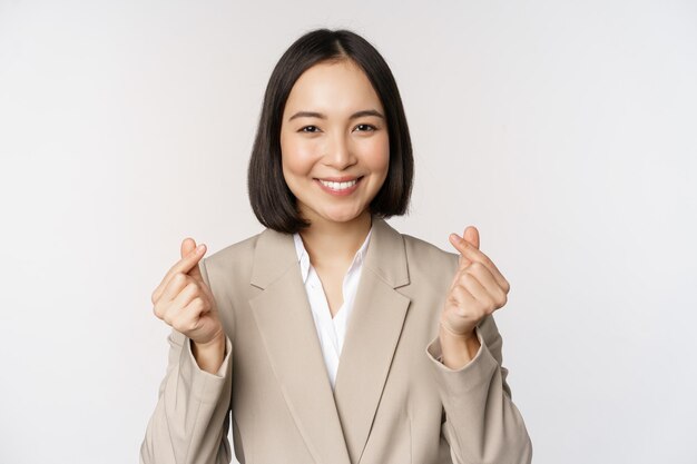 Cheerful asian saleswoman smiling and showing finger hearts sign standing in suit over white background