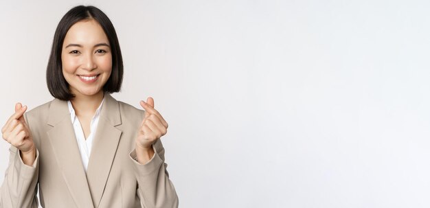 Cheerful asian saleswoman smiling and showing finger hearts sign standing in suit over white background