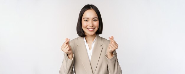 Cheerful asian saleswoman smiling and showing finger hearts sign standing in suit over white background