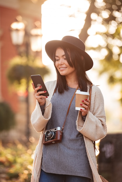 검은 모자와 거리에서 커피를 마시는 동안 휴대 전화에 뉴스를 확인 모직 카디건에 쾌활 한 아시아 여자