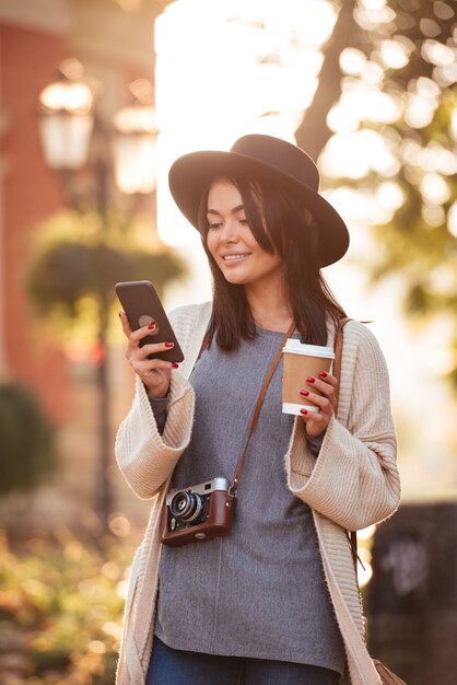 Жизнерадостная азиатская женщина в черной шляпе и шерстяной кардиган, проверка новостей на мобильном телефоне, попивая кофе на улице