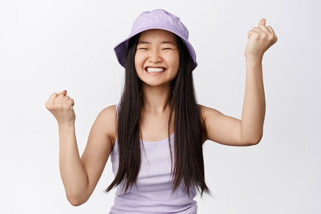 쾌활한 아시아 여성이 축하하고, 주먹을 쥔 채 만족스럽게 웃고, 기쁨에서 승리하고, 흰색 위에 서 있습니다.